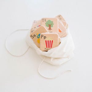 May include: A white fabric bag filled with wooden alphabet blocks. The blocks have letters and pictures on them, including a tree, a car, and popcorn.