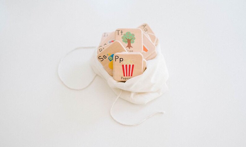 May include: A white fabric bag filled with wooden alphabet blocks. The blocks have letters and pictures on them, including a tree, a car, and popcorn.