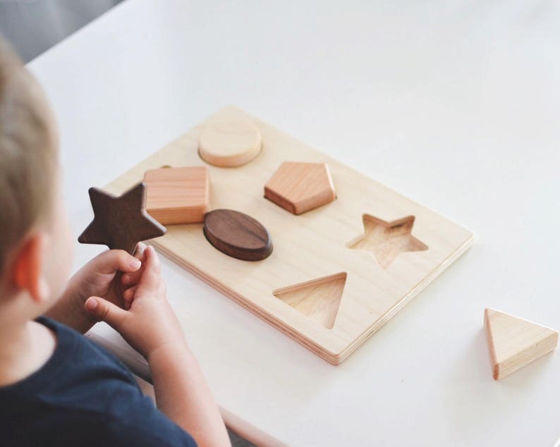 Puede incluir: Rompecabezas de formas de madera con una base de madera clara y varias formas, incluyendo una estrella, un c&iacute;rculo y un tri&aacute;ngulo. Las piezas del rompecabezas son en tonos de madera natural, con una estrella marr&oacute;n oscuro. Juguete educativo para ni&ntilde;os peque&ntilde;os.