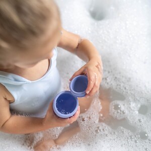 Puede incluir: Un ni&ntilde;o peque&ntilde;o con un traje de ba&ntilde;o blanco se sienta en una ba&ntilde;era llena de burbujas, sosteniendo dos juguetes de ba&ntilde;o morados.