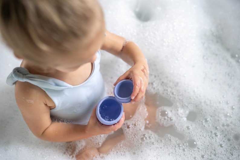 Puede incluir: Un ni&ntilde;o peque&ntilde;o con un traje de ba&ntilde;o blanco se sienta en una ba&ntilde;era llena de burbujas, sosteniendo dos juguetes de ba&ntilde;o morados.