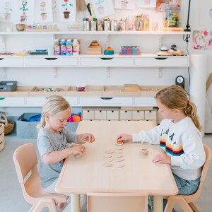 May include: Two young girls sit at a light wood table, playing with wooden discs. The room is a bright, well-lit space with white shelves filled with art supplies and drawings. The girls are wearing casual clothing.