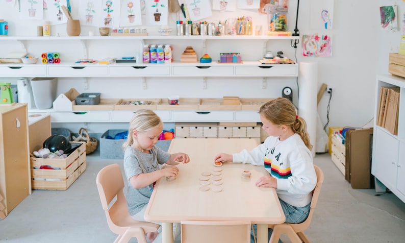 May include: Two young girls sit at a light wood table, playing with wooden discs. The room is a bright, well-lit space with white shelves filled with art supplies and drawings. The girls are wearing casual clothing.