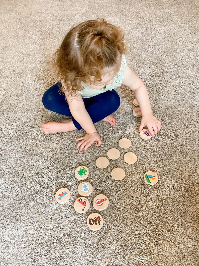 May include: A child is playing with wooden circle toys with colorful illustrations. The child is wearing a light green top and blue pants. The toys are scattered on a beige carpet.