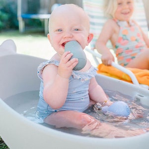 Puede incluir: Un beb&eacute; con un traje de ba&ntilde;o a rayas azul y blanco se sienta en una ba&ntilde;era blanca con forma de ballena llena de agua. El beb&eacute; sostiene un juguete gris en la boca y sonr&iacute;e.
