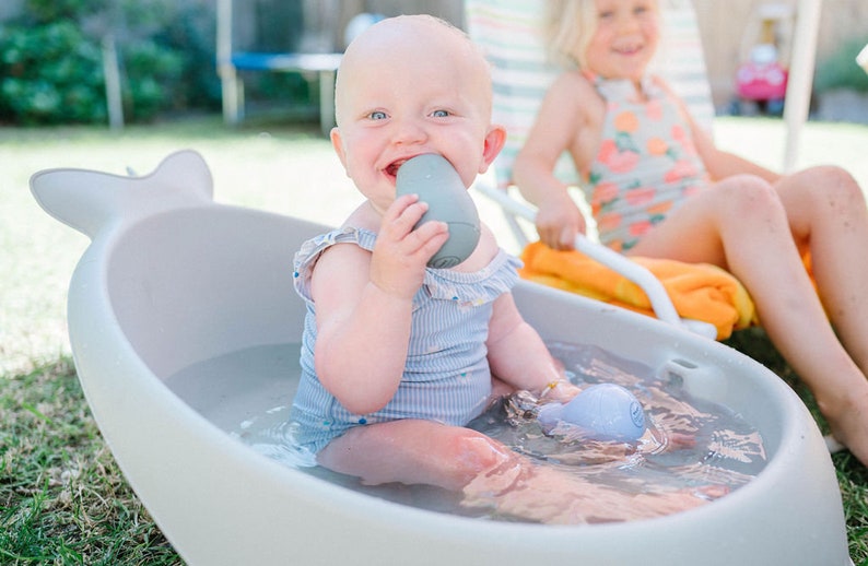 Puede incluir: Un beb&eacute; con un traje de ba&ntilde;o a rayas azul y blanco se sienta en una ba&ntilde;era blanca con forma de ballena llena de agua. El beb&eacute; sostiene un juguete gris en la boca y sonr&iacute;e.