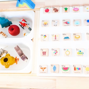 May include: A white board with colorful alphabet letter tiles featuring illustrations of various objects. Each tile has a letter and a word describing the image. The letters are in alphabetical order from A to Z. The illustrations include an apple, bear, cactus, donut, egg, fire, goat, helicopter, igloo, jellyfish, kite, lighthouse, mountains, nest, otter, popcorn, quilt, ring, submarine, tree, underwear, vase, watermelon, xylophone, yarn, and zebra.