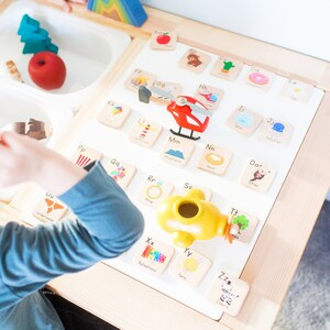May include: A wooden table with a white surface featuring alphabet letter tiles with corresponding images. The tiles are arranged in alphabetical order. A red toy helicopter is on the table.