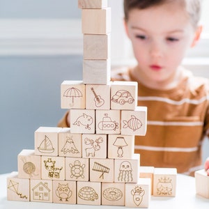 May include: Wooden alphabet blocks stacked in a tower. Each block features a different etched image, including an umbrella, guitar, car, whale, submarine, and fish. The blocks are light brown and arranged on a white surface.