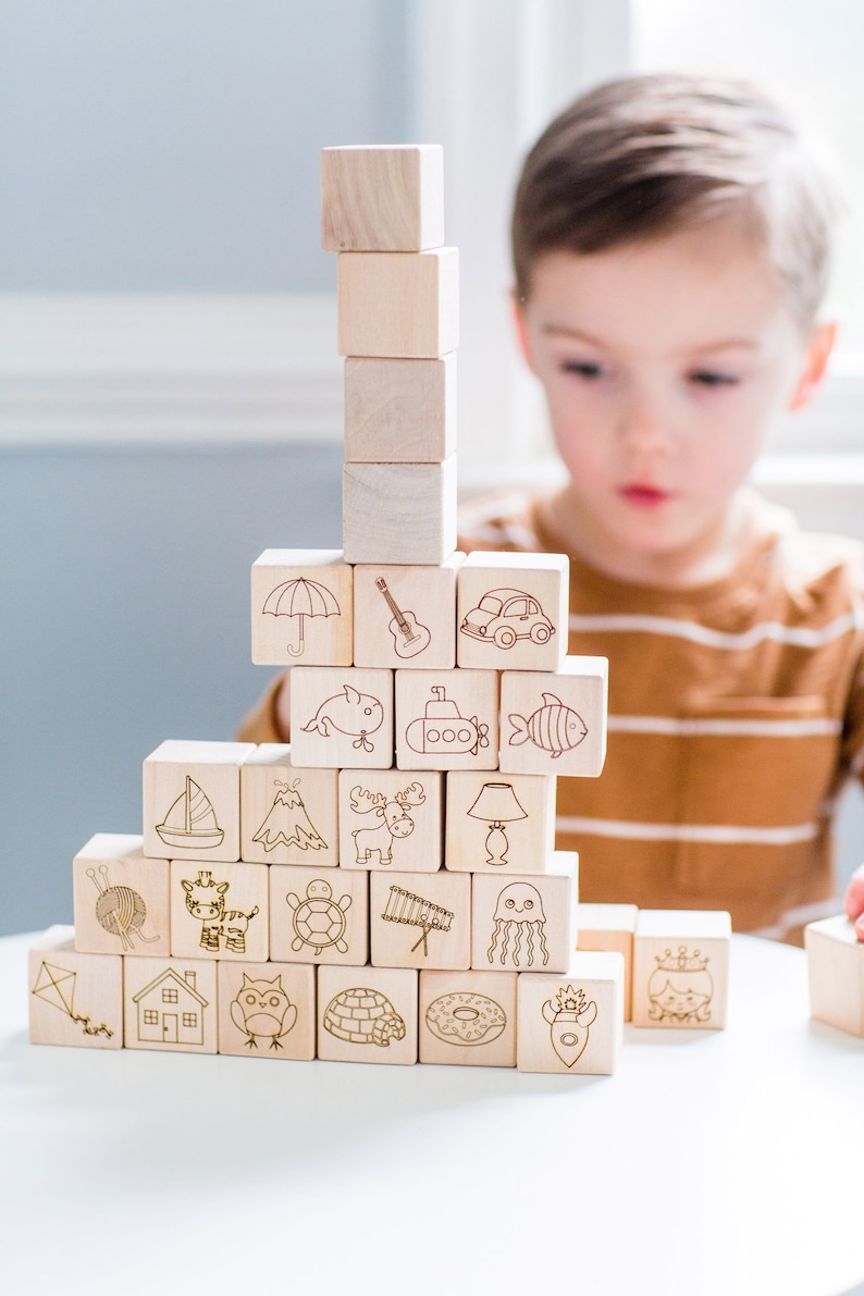 May include: Wooden alphabet blocks stacked in a tower. Each block features a different etched image, including an umbrella, guitar, car, whale, submarine, and fish. The blocks are light brown and arranged on a white surface.