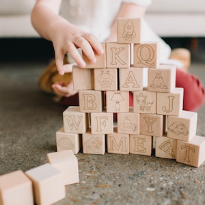 May include: Wooden alphabet blocks with engraved letters and illustrations, including a rocket, owl, and car. The blocks are stacked in a tower, with a child's hand reaching towards them. The blocks are made of light-colored wood.