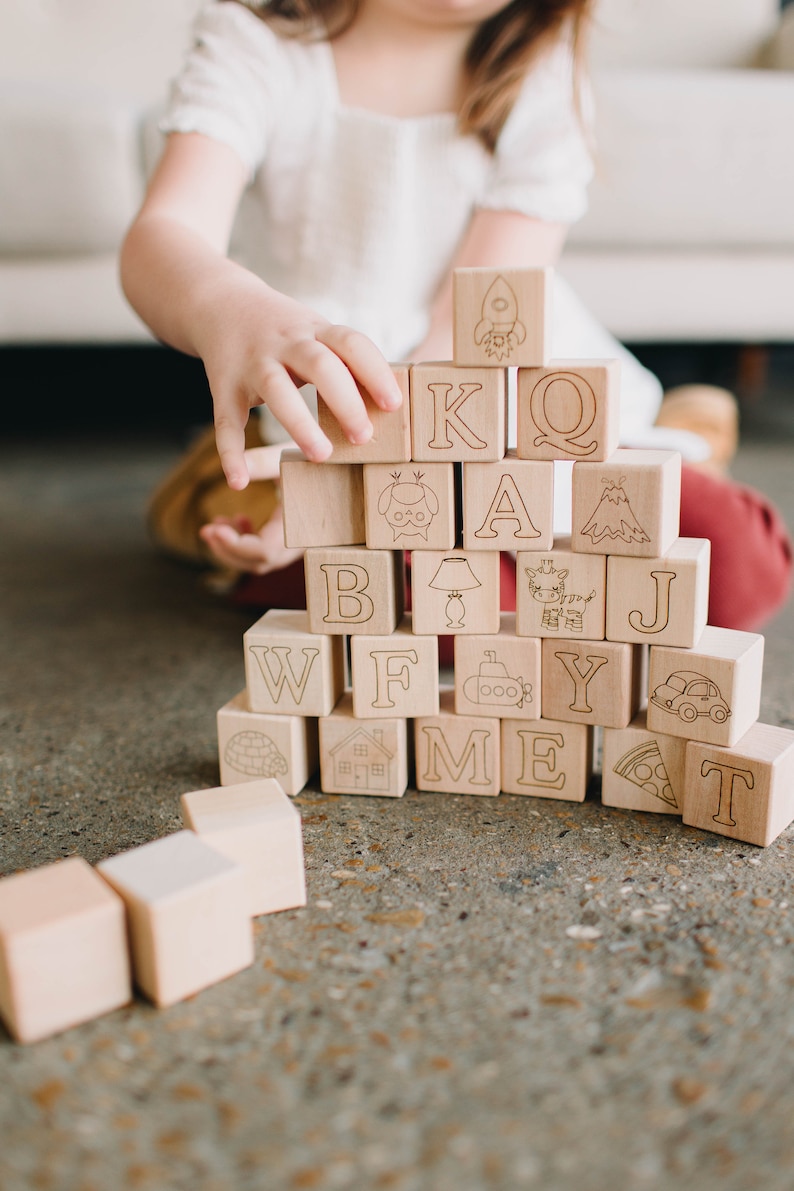 May include: Wooden alphabet blocks with engraved letters and illustrations, including a rocket, owl, and car. The blocks are stacked in a tower, with a child's hand reaching towards them. The blocks are made of light-colored wood.