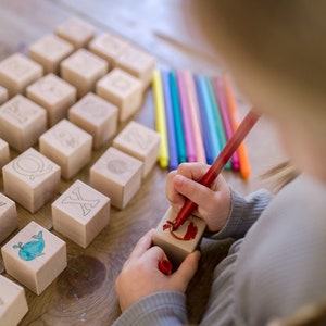 May include: Wooden alphabet blocks with engraved letters and illustrations, including a pizza slice and a whale. A child is using a red marker to color a block. Colored markers are arranged nearby on a wooden surface. Educational toy.