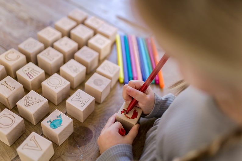 May include: Wooden alphabet blocks with engraved letters and illustrations, including a pizza slice and a whale. A child is using a red marker to color a block. Colored markers are arranged nearby on a wooden surface. Educational toy.