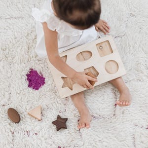 Puede incluir: Un ni&ntilde;o juega con un clasificador de formas de madera sobre una alfombra blanca y esponjosa. El clasificador tiene recortes de diferentes formas, y el ni&ntilde;o sostiene una forma de madera. Otras formas de madera est&aacute;n dispersas.
