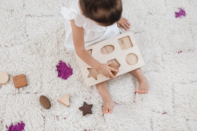 Puede incluir: Un ni&ntilde;o juega con un clasificador de formas de madera sobre una alfombra blanca y esponjosa. El clasificador tiene recortes de diferentes formas, y el ni&ntilde;o sostiene una forma de madera. Otras formas de madera est&aacute;n dispersas.