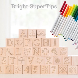 May include: Wooden alphabet blocks with etched letters, numbers, and illustrations, stacked in a pyramid. A set of colorful markers with white barrels and colored tips are arranged diagonally. The text "Bright SuperTips" is at the top.