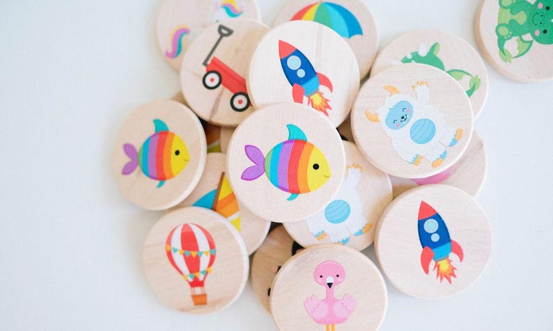 May include: Wooden circle game pieces with colorful illustrations of a fish, rocket, hot air balloon, and other whimsical designs. The pieces are scattered on a white surface, creating a playful and engaging visual.