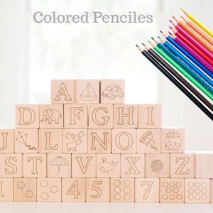 May include: Wooden alphabet blocks with etched letters and illustrations, stacked in a pyramid shape. A set of colored pencils in various colors is positioned to the right. The text "Colored Pencils" is at the top.