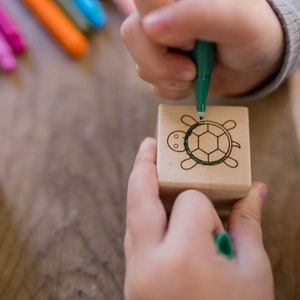 May include: A child's hands drawing a green turtle on a wooden block with a green marker. Other colorful markers are visible in the background. The block has a turtle design etched into it.