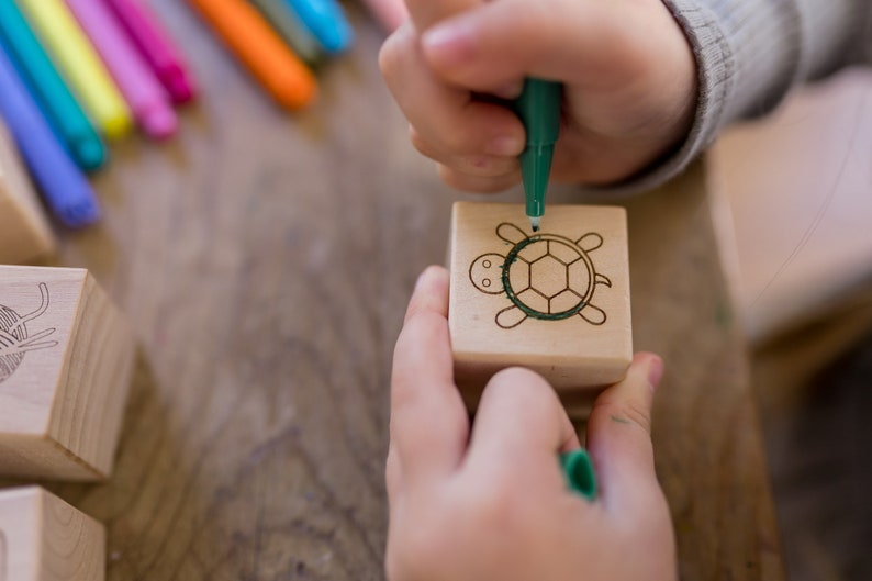 May include: A child's hands drawing a green turtle on a wooden block with a green marker. Other colorful markers are visible in the background. The block has a turtle design etched into it.