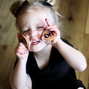 May include: A young child holds two wooden circles with the word "off" in black letters. The child is wearing a black shirt and has their hair up in two small buns. The background is a wooden floor.