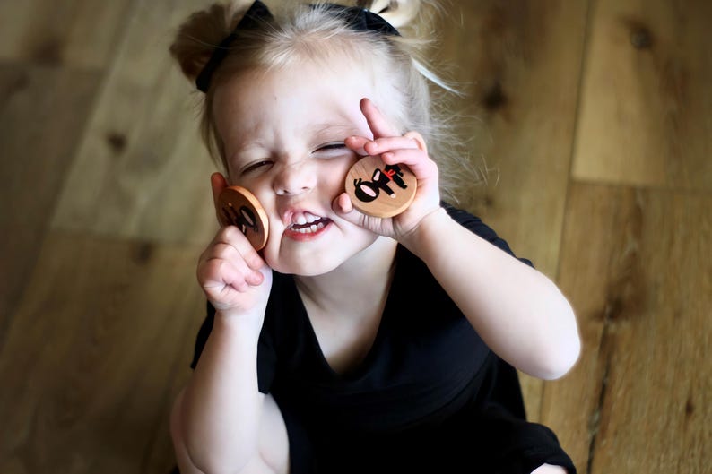 May include: A young child holds two wooden circles with the word "off" in black letters. The child is wearing a black shirt and has their hair up in two small buns. The background is a wooden floor.