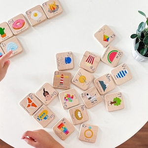 May include: Wooden alphabet blocks with colorful illustrations and corresponding letters. Each block features a different object, such as an apple, bear, cactus, donut, egg, fire, helicopter, ice cream, lighthouse, mountain, ring, tree, and zebra. The blocks are arranged in a circle on a white table.