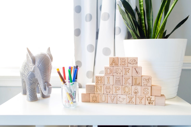 May include: A collection of wooden alphabet blocks with engraved letters and illustrations, arranged in a pyramid. A gray elephant figurine and a glass jar of colorful markers sit nearby. A potted plant and a polka-dot curtain are in the background.