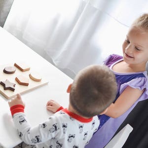 Puede incluir: Un rompecabezas de clasificaci&oacute;n de formas de madera con varias formas geom&eacute;tricas, incluyendo una estrella, un hex&aacute;gono y un rect&aacute;ngulo. El rompecabezas est&aacute; sobre una base de madera de color claro. Dos ni&ntilde;os interact&uacute;an con el rompecabezas, uno se&ntilde;al&aacute;ndolo.