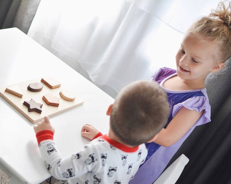 Puede incluir: Un rompecabezas de clasificaci&oacute;n de formas de madera con varias formas geom&eacute;tricas, incluyendo una estrella, un hex&aacute;gono y un rect&aacute;ngulo. El rompecabezas est&aacute; sobre una base de madera de color claro. Dos ni&ntilde;os interact&uacute;an con el rompecabezas, uno se&ntilde;al&aacute;ndolo.