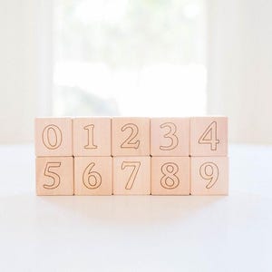 May include: Ten wooden blocks, each displaying a number from 0 to 9. The numbers are etched in a dark color on the light-colored wood. The blocks are arranged in two rows of five, sitting on a white surface.
