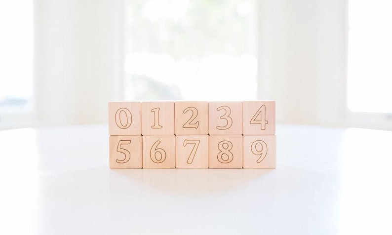 May include: Ten wooden blocks, each displaying a number from 0 to 9. The numbers are etched in a dark color on the light-colored wood. The blocks are arranged in two rows of five, sitting on a white surface.