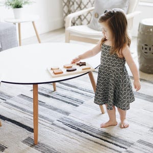 Puede incluir: Una mesa blanca con tapa redonda y patas de madera. Una bandeja de rompecabezas de madera con varias formas est&aacute; sobre la mesa. Un ni&ntilde;o con un vestido estampado en blanco y negro est&aacute; de pie cerca de la mesa.