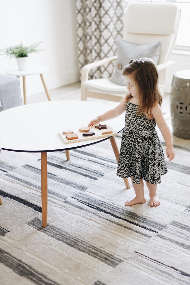 Puede incluir: Una mesa blanca con tapa redonda y patas de madera. Una bandeja de rompecabezas de madera con varias formas est&aacute; sobre la mesa. Un ni&ntilde;o con un vestido estampado en blanco y negro est&aacute; de pie cerca de la mesa.