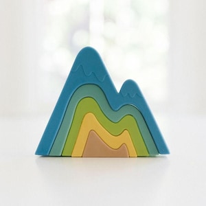 May include: A mountain-shaped stacking toy consisting of five nested pieces in a gradient of colors, including blue, teal, light green, yellow, and tan. Each piece features a smooth, curved design that fits together to form a single peak.