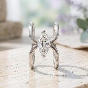 Peut inclure: Une bague argentée dotée d'une pierre précieuse transparente centrale de taille marquise, maintenue entre deux bandes métalliques incurvées et parallèles qui s'étendent depuis la pierre, créant un design architectural ouvert et poli.