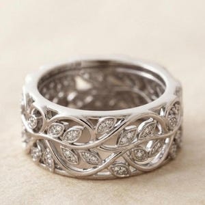 Puede incluir: Un anillo de metal plateado con un diseño intrincado y calado de enredaderas y hojas interconectadas. La superficie de las hojas está adornada con pequeñas piedras claras y brillantes incrustadas a lo largo de la banda, creando un patrón continuo.