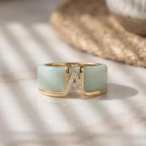 Peut inclure: Une bague dorée avec une bande vert clair et la lettre "A" ornée de petites pierres transparentes. La bague présente un design géométrique moderne et est posée sur un fond doux et neutre. Il s'agit d'un bijou.