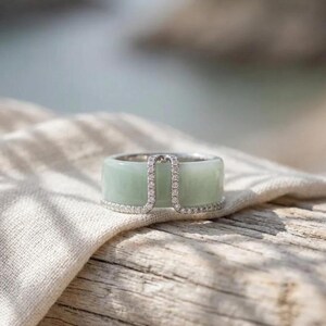 May include: A wide band ring with a pale green jade centre and a silver-coloured frame. The frame is embellished with small, clear gemstones. The ring is shown on a light-coloured fabric and a wooden surface.