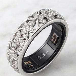 Puede incluir: Un anillo inteligente de tono plateado con un intrincado patrón floral calado incrustado con pequeñas piedras transparentes. El interior de la banda es negro con el texto OURA impreso en blanco e incluye componentes de sensores electrónicos.