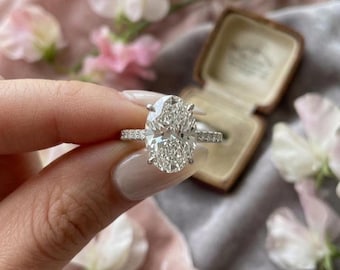May include: A diamond engagement ring with an oval-cut center stone and a pavé band. The ring is held up against a backdrop of flowers and a vintage ring box. The ring's center stone is a brilliant white color.