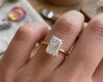 May include: A radiant-cut clear gemstone ring set on a thin, polished yellow gold band. The rectangular stone features multiple facets that reflect light, held in place by four delicate gold prongs. The band is simple, smooth, and circular.