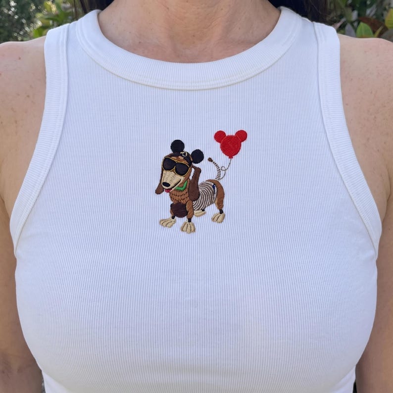 May include: White ribbed tank top with an embroidered cartoon dachshund wearing sunglasses and a Mickey Mouse-shaped balloon. The dachshund has a brown body, a skeleton-like design, and is holding a red balloon.