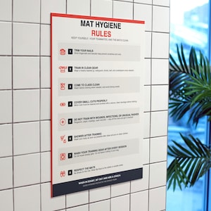 Op de afbeelding: Een rechthoekig bord met de tekst "MAT HYGIENE RULES" in rood en zwart. Het bord geeft acht regels voor het handhaven van de hygiëne, waaronder het knippen van nagels, het dragen van schone kleding en het douchen na de training. Het bord is bevestigd aan een wit betegelde muur.