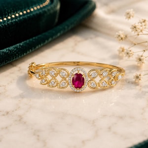 Puede incluir: Un brazalete rígido de oro con bisagra que presenta una piedra preciosa roja oscura de corte ovalado en el centro, rodeada por un halo de pequeñas piedras blancas. La banda tiene motivos simétricos en forma de hoja con piedras blancas.