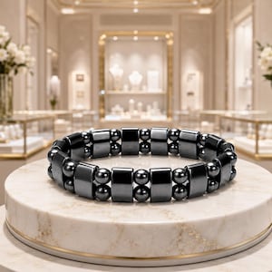 Könnte beinhalten: Ein zweireihiges Armband mit abwechselnd dunkelgrauen, polierten zylindrischen Perlen und Paaren kleinerer, kugelförmiger Perlen. Das Armband hat eine glatte, reflektierende metallische Oberfläche und ist in einer durchgehenden Kreisform angeordnet.