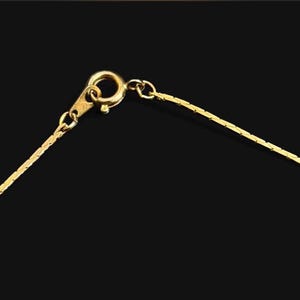May include: Close-up of a gold-colored necklace against a black background. The necklace features a delicate chain and a lobster clasp. The chain is made of small, rectangular links that catch the light.