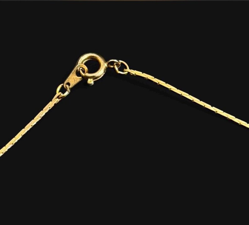 May include: Close-up of a gold-colored necklace against a black background. The necklace features a delicate chain and a lobster clasp. The chain is made of small, rectangular links that catch the light.