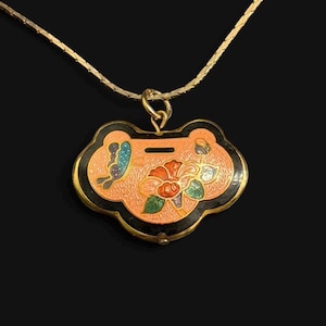 May include: A gold-toned necklace with a pendant featuring a floral design. The pendant has a peach-colored background with a black border and a butterfly and flower design. The necklace is on a black background.